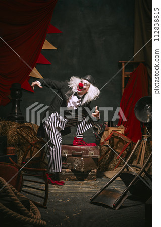Clown in black hat, white face with red nose and striped pants playing over dark retro circus backstage background. Comedian Clown in black hat, white face with red nose and striped pants playing over dark retro circus backstage background. Comedian 112838815