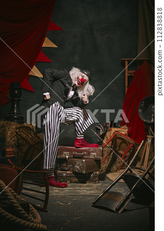 Clown emotional man in black hat, white face with red nose and striped pants playing over dark retro circus backstage background. Clown emotional man in black hat, white face with red nose and striped pants playing over dark retro circus backstage background. 112838818