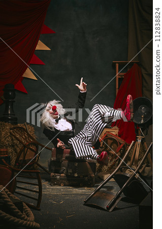 Clown in black hat, white face with red nose and striped pants playing with emotive face over dark retro circus backstage background. Clown in black hat, white face with red nose and striped pants playing with emotive face over dark retro circus backstage background. 112838824