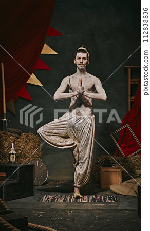 Meditation performance. Artistic image of pale man, shirtless yogi performing over dark retro circus backstage background. Meditation performance. Artistic image of pale man, shirtless yogi performing over dark retro circus backstage background. 112838836
