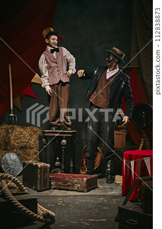 Little servant boy standing on chair near giant African man with dirty face and clothes over dark retro circus backstage background. 112838873