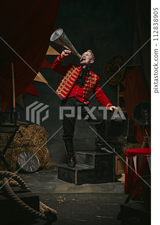 Announcement. Poster. Ringmaster, magician in red coat shouting into megaphone over dark retro circus backstage background. Announcement. Poster. Ringmaster, magician in red coat shouting into megaphone over dark retro circus backstage background. 112838905