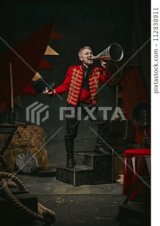 Ringmaster, magician in red coat shouting into megaphone over dark retro circus backstage background. Announcement of show 112838911