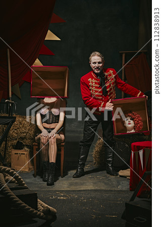 Tricks. Magician in red coat showing tricks with headless female assistant holding boxes over dark retro circus backstage background. Tricks. Magician in red coat showing tricks with headless female assistant holding boxes over dark retro circus backstage background. 112838913
