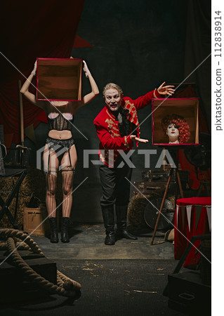 Tricks. Magician in red coat showing tricks with headless female assistant holding boxes over dark retro circus backstage background. 112838914