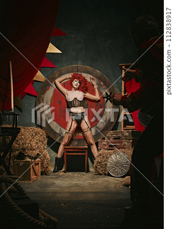 Young redhead woman with stage makeup and costume near target over dark retro circus backstage background. Man throwing knives 112838917