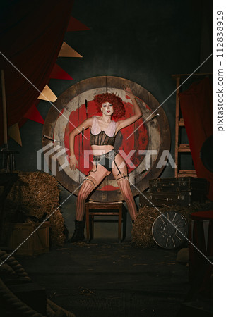 Woman in vintage costume and stage makeup poses before target over dark retro circus backstage background. Dangerous tricks 112838919