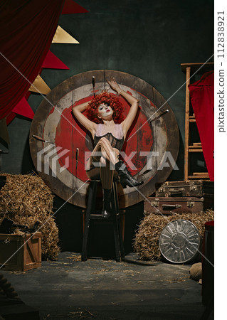 Danger. Elegant young woman with bright makeup and stage costume sitting next to target with knives over dark retro circus backstage background. 112838921