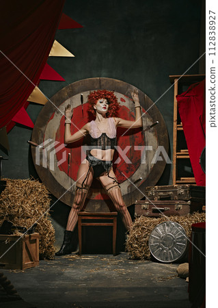 Young redhead woman with stage makeup and costume standing near target over dark retro circus backstage background. 112838927