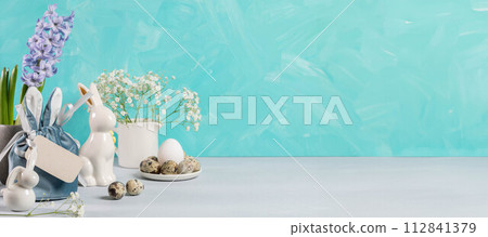 Easter banner. Eggs, ceramic bunnies, flowers, gift bag, blank note on turquoise. Copy space. Easter banner. Eggs, ceramic bunnies, flowers, gift bag, blank note on turquoise. Copy space. 112841379