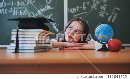 Adorable school girl posing at her desk against a background of blackboard, books, globe and graduation cap. 112841715