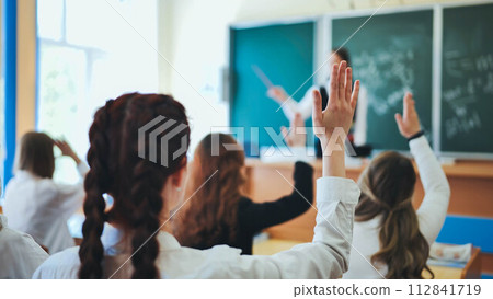 Intelligent group of young school children all raising their hands in the air to answer a question posed by the female teacher, view from behind Intelligent group of young school children all raising their hands in the air to answer a question posed by the female teacher, view from behind 112841719