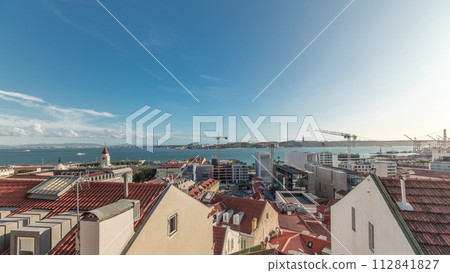 Panorama showing red roofs timelapse and 25 de Abril Bridge, Iconic suspension bridge over Tagus River in Lisbon, Portugal. 112841827