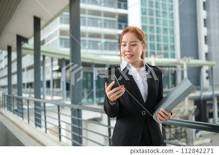 A young Asian businesswomen wearing a suit holding files standing in a big city on a busy downtown street. Young Asian businesswoman using smartphone texting to contact clients. 112842271