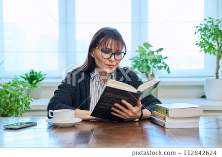 Middle-aged woman reading books, sitting at table at home Middle-aged woman reading books, sitting at table at home 112842624