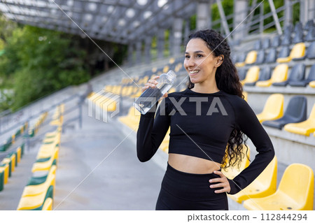 A fitness enthusiast holding a water bottle, taking a break during an outdoor workout session in a stadium. 112844294