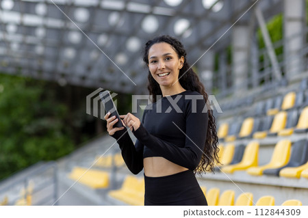 A smiling young woman wearing athletic wear holds a smartphone while standing at a sports stadium with seats in the background. 112844309