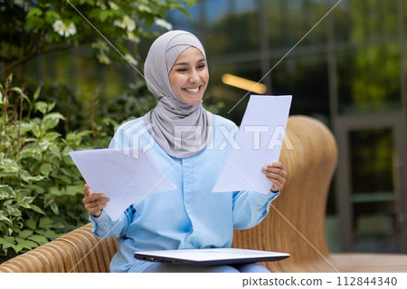 A cheerful Muslim woman in a hijab holding documents, smiling outdoors with a blurred green background, portraying confidence and positivity. A cheerful Muslim woman in a hijab holding documents, smiling outdoors with a blurred green background, portraying confidence and positivity. 112844340