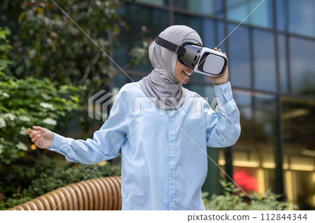 Smiling Muslim lady in hijab using virtual reality headset outside, experiencing modern technology in a natural setting. 112844344