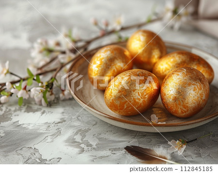 Festive Easter painted golden eggs at the ceramic plate with small feathers on a light grey background 112845185