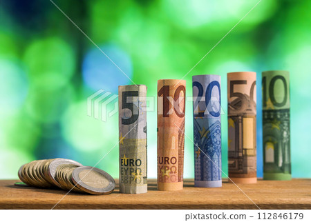 Five, ten, twenty, fifty and one hundred euro rolled bills banknotes, with euro coins on green blurred bokeh background. Five, ten, twenty, fifty and one hundred euro rolled bills banknotes, with euro coins on green blurred bokeh background. 112846179