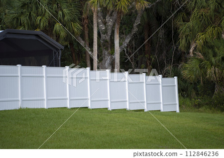White vinyl picket fence on green lawn surrounding property grounds for backyard protection and privacy White vinyl picket fence on green lawn surrounding property grounds for backyard protection and privacy 112846236