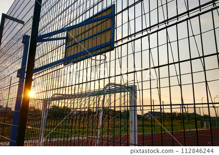 Outdoors mini football and basketball court with ball gate and basket surrounded with high protective fence. 112846244
