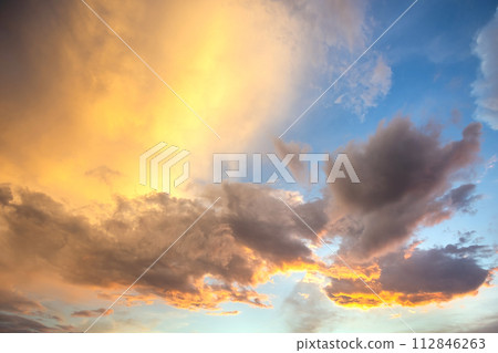 Dramatic sunset landscape with puffy clouds lit by orange setting sun and blue sky. 112846263