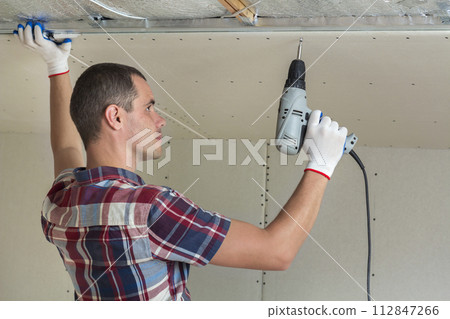 Young man in usual clothing and work gloves fixing drywall suspended ceiling to metal frame using electrical screwdriver on ceiling insulated with shiny aluminum foil. DIY, do it yourself concept. 112847266
