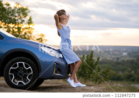 Young woman driver enjoying warm summer day standing beside her car. Travelling and vacation concept. 112847275
