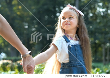 Young mom and her small daughter with long hair walking together holding hands in summer park. 112847279