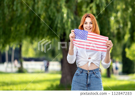 Happy young woman posing with USA national flag outdoors in summer park. Pretty girl celebrating United States independence day. 112847283