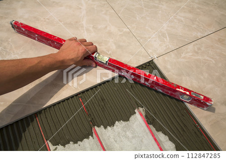 Ceramic tiles and tools for tiler. Worker hand installing floor tiles. Home improvement, renovation - ceramic tile floor adhesive, mortar, level. Ceramic tiles and tools for tiler. Worker hand installing floor tiles. Home improvement, renovation - ceramic tile floor adhesive, mortar, level. 112847285