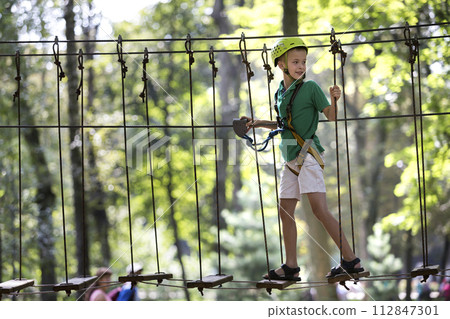 Young child boy in safety harness and helmet attached with carbine to cable on rope way in park. Young child boy in safety harness and helmet attached with carbine to cable on rope way in park. 112847301