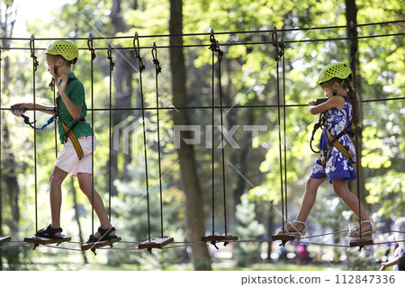 Two children, boy and girl in protective harness and safety helmets at climbing activity on rope way. 112847336