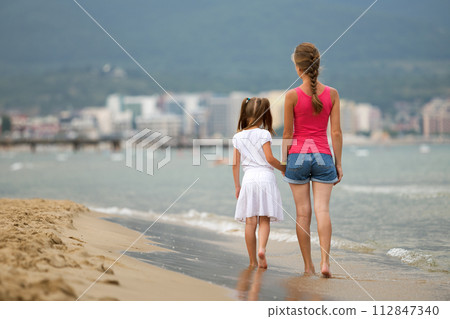 Mother and daughter girl walking together on sand beach in sea water in summer with bare feet in warm ocean waves. 112847340