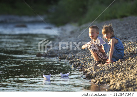 Two cute blond children, boy and girl on river bank sending in water white paper boats on bright summer blurred blue background. Joys and games of happy childhood and outdoors activities concept. Two cute blond children, boy and girl on river bank sending in water white paper boats on bright summer blurred blue background. Joys and games of happy childhood and outdoors activities concept. 112847347