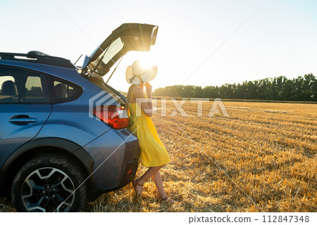 Young woman driver in yellow summer dress and straw hat standing near her car enjoying warm summer day at sunset. Young woman driver in yellow summer dress and straw hat standing near her car enjoying warm summer day at sunset. 112847348