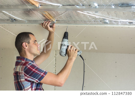 Young man in usual clothing and work gloves fixing drywall suspended ceiling to metal frame using electrical screwdriver on ceiling insulated with shiny aluminum foil. DIY, do it yourself concept. Young man in usual clothing and work gloves fixing drywall suspended ceiling to metal frame using electrical screwdriver on ceiling insulated with shiny aluminum foil. DIY, do it yourself concept. 112847380