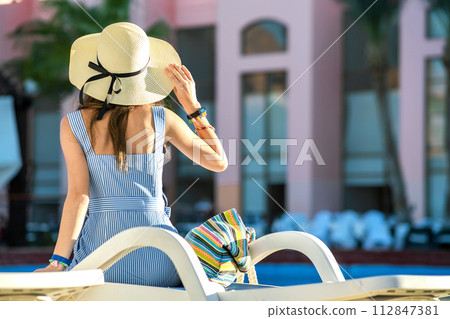 Young woman wearing light summer dress and yellow straw hat sitting outside near hotel swimming pool on summer sunny day. Young woman wearing light summer dress and yellow straw hat sitting outside near hotel swimming pool on summer sunny day. 112847381