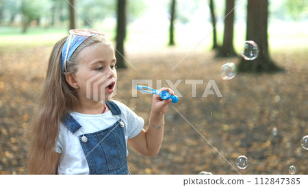 Little happy child girl blowing soap bubbles outside in green park. Outdoor summer activities concept. Little happy child girl blowing soap bubbles outside in green park. Outdoor summer activities concept. 112847385
