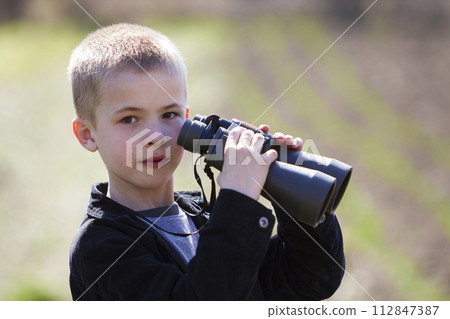 Portrait in profile of little handsome cute blond boy looking thoughtfully through binoculars in distance on blurred background. Children dreams, fantasies and imaginations concept. Portrait in profile of little handsome cute blond boy looking thoughtfully through binoculars in distance on blurred background. Children dreams, fantasies and imaginations concept. 112847387