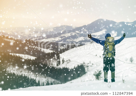 Back view of tourist hiker in warm clothing with backpack standing with raised arms on mountain clearing on copy space background of woody mountain ridge and cloudy sky. Back view of tourist hiker in warm clothing with backpack standing with raised arms on mountain clearing on copy space background of woody mountain ridge and cloudy sky. 112847394