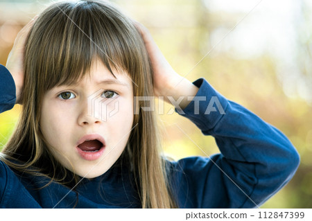 Portrait of surprised child girl holding hands to her head outdoors in summer. Shocked female kid on a warm day outside. Portrait of surprised child girl holding hands to her head outdoors in summer. Shocked female kid on a warm day outside. 112847399