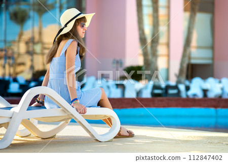 Young woman wearing light summer dress and yellow straw hat sitting outside near hotel swimming pool on summer sunny day. 112847402