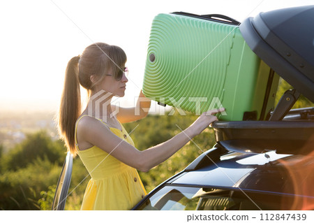 Female driver putting green suitcase inside her car roof trunk. Travel and vacations concept. 112847439