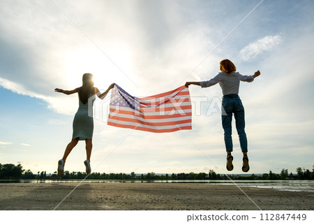 Two young friends women holding USA national flag jumping up together outdoors at sunset. Patriotic girls celebrating United States independence day. 112847449