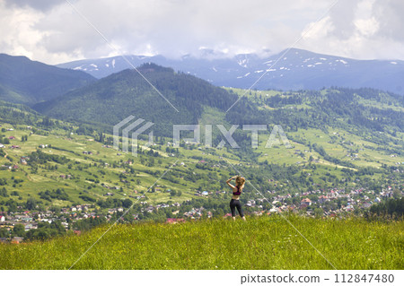 Slim young woman with raised arms outdoors on background of beautiful mountain landscape on sunny summer day. 112847480