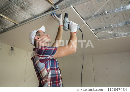 Young man in usual clothing and work gloves fixing drywall suspended ceiling to metal frame using electrical screwdriver on ceiling insulated with shiny aluminum foil. DIY, do it yourself concept. 112847484