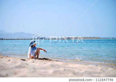Lonely child girl playing with white sand on sea beach on background of blue sky, clear water and distant shore. Summer tourism and recreation concept Lonely child girl playing with white sand on sea beach on background of blue sky, clear water and distant shore. Summer tourism and recreation concept 112847746